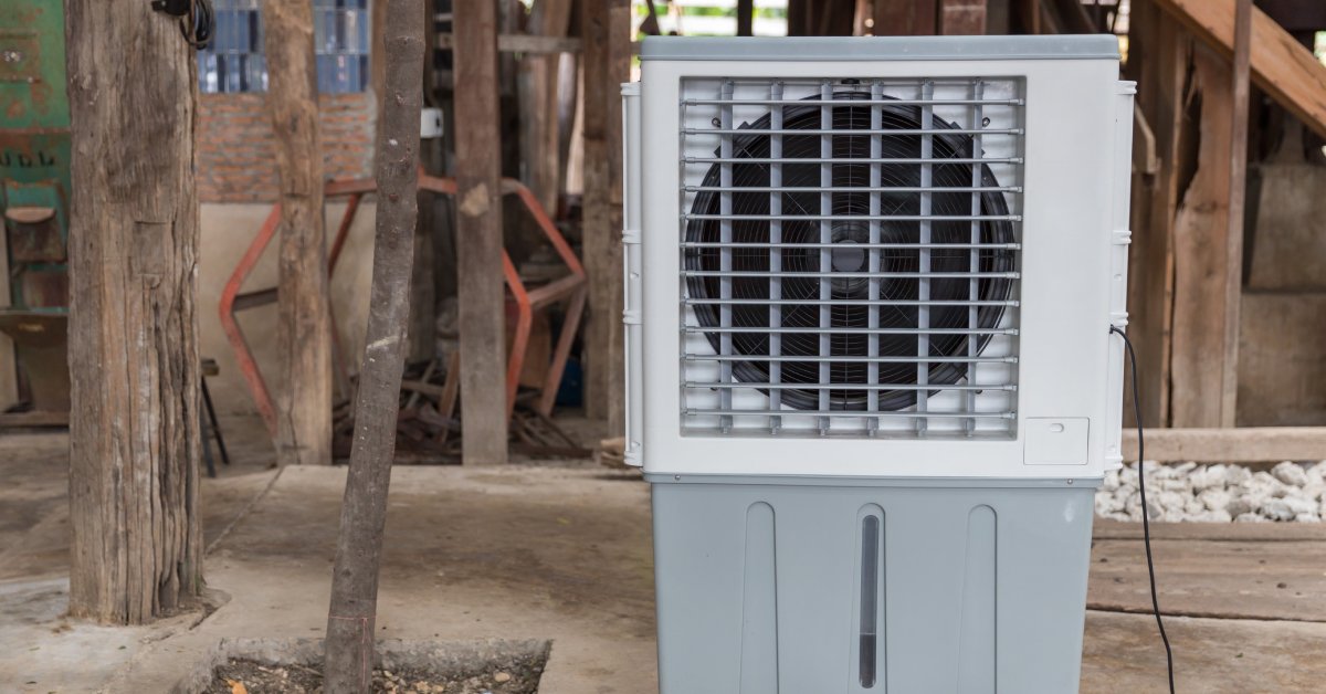 A portable evaporative air cooling fan sits on a cement surface. Wood, rocks, and miscellaneous things are behind the cooler.